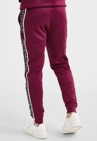 Burgundy joggers med en svart och vit rand längs sömmen som har texten "MESSISILK", avslutade med elastiska manschetter och en slät tyg.
