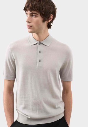 Młody mężczyzna w jasnoszarej koszulce polo z krótkim rękawem, patrzący w lewo, na jednolitym jasnym tle.