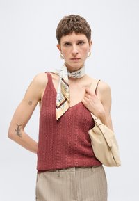 Γυναίκα με κοντά μαλλιά φοράει μια υφήτο, σκουριασμένου χρώματος αμάνικο τοπ, κασκόλ με σχέδιο, μπεζ παντελόνι και κρατά μια μπεζ τσάντα ώμου, ουδέτερο φόντο.