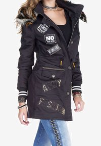 Giacca lunga nera con cappuccio foderato in pelliccia, caratterizzata da vari toppi, borchie e "ARMY FASHION" in strass. Polsini e tasche a righe.