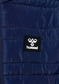 Granatowa puchowa kurtka z pikowanym wzorem, z czarną naszywką z logo z przodu. Gładka tekstura z lekkiego materiału.