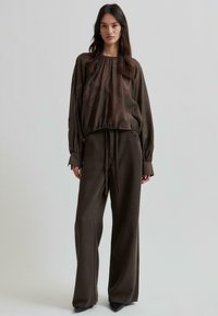 Blouse légère marron avec broderie florale, col froncé, manches longues ; associée à un pantalon large assorti et des chaussures noires à talons.
