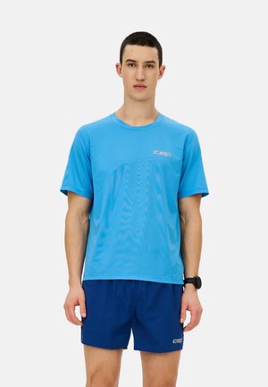 Junger Mann, der ein hellblaues Sport-T-Shirt und dunkelblaue Shorts trägt, steht mit Blick nach vorne vor einem einfachen Hintergrund.
