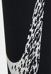 Černé legíny s velkým bílým vzorem ve tvaru swoosh s texturovaným, rozpraskaným designem. Tkanina má hladký, elastický vzhled.