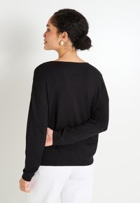 Mujer con cabello rizado oscuro recogido en un moño, que lleva pendientes en forma de lágrima dorados, una blusa negra de manga larga y pantalones blancos, de pie contra un fondo blanco.