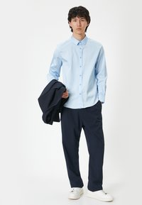 Ljusblå skjorta med långa ärmar, kombinerad med mörkblå byxor och hållande en matchande jacka, bärs med vita sneakers.