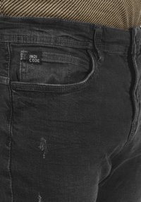 Jeans de mezclilla negra con un ajuste relajado, un leve desgaste y una etiqueta con "INDI CODE" en el bolsillo trasero. Tejido texturizado.