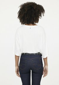 Weißes, locker sitzendes Oberteil mit kurzen Ärmeln, kombiniert mit dunklen Denim-Jeans mit orangefarbenen Nähten und einem dekorativen Nieten-Gürtel.