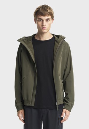 Maslinasto zelena jakna od softshell materijala s prilagođenom kapom, zatvaračem, elastičnim manžetama i dva bočna džepa, nošena preko crne majice.
