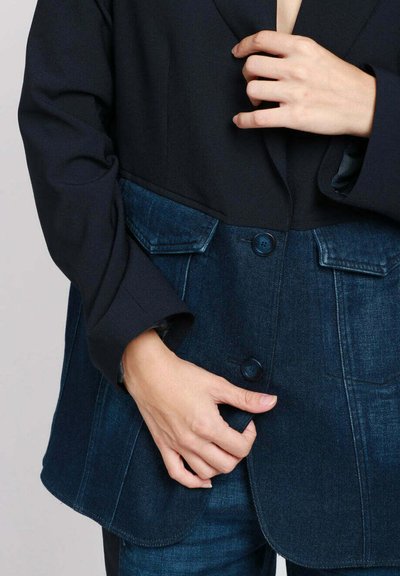 Persona che indossa una giacca di jeans nera e blu scura, mentre abbottona la parte frontale, mostrando le tasche e le maniche della giacca.