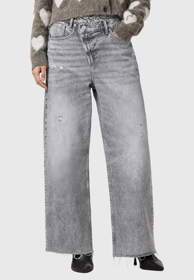 Jeans grigi ampi con un design strappato, con orli sfilacciati e un leggero scolorimento. Indossati con un maglione a fantasia e scarpe nere.