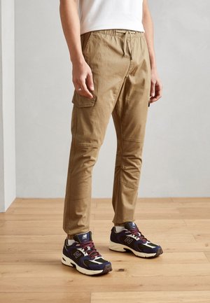 Osoba nosząca beżowe spodnie cargo ze ściągaczem w talii i granatowe trampki New Balance stoi na drewnianej podłodze.