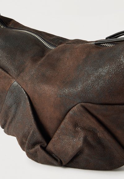 Μαλακή, φθαρμένη δερμάτινη τσάντα duffel σκούρου καφέ χρώματος με εμφανές ασημί φερμουάρ και υφιστάμενη υφή, εμφανιζόμενη πάνω σε απλό φόντο.