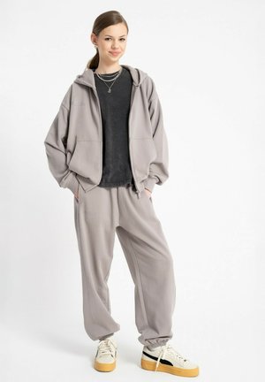 Młoda kobieta stoi z rękami w kieszeniach, ubrana w jasnoszary dres, czarną koszulkę, warstwowe naszyjniki i białe sneakersy z żółtymi podeszwami.