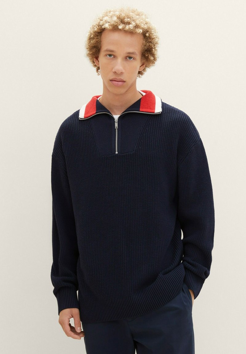 Marineblauer Strickpullover mit einem halben Reißverschluss und roten sowie weißen Akzenten. Lockerer Schnitt mit gerippten Bündchen und Saum, strukturierter Stoff.
