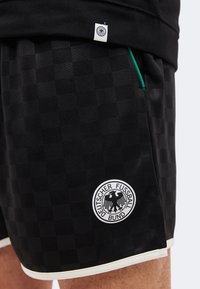 Μαύρο καρό αθλητικό σορτς με το λογότυπο της Deutscher Fußball-Bund, λευκή επένδυση και πράσινη λεπτομέρεια στην άκρη της τσέπης στον μηρό ενός ατόμου.