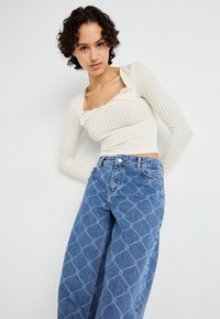 Off-white ribbed long-sleeve top with ruffled neckline paired with blue denim wide-leg jeans featuring a geometric print.