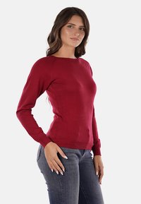 Maglione rosso con maniche lunghe realizzato in tessuto morbido, caratterizzato da un neckline rotondo e un design aderente, abbinato a jeans blu scuro.