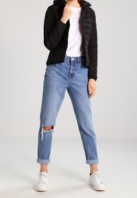 Schwarze gesteppte Jacke mit Kapuze, weißes T-Shirt und hellblaue zerrissene Jeans. Weiße Sneakers vervollständigen das Outfit.