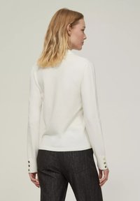 Top bianco a maniche lunghe con un design aderente, texture a coste e dettagli con bottoni sulle maniche, abbinato a pantaloni scuri grigio con motivo.