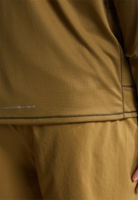 Brązowy, teksturowany sportowy top z wzorem w kratę; posiada zakrzywiony dół i kontrastujące przeszycia. Połączony z jednolitymi brązowymi spodami.