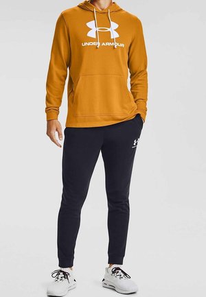Homme portant un sweat à capuche jaune moutarde Under Armour, un jogging noir Under Armour et des chaussures de sport blanches, debout devant un fond uni.