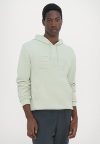 Felpa con cappuccio verde menta caratterizzata da un logo "EA7" testurizzato, tasca a marsupio e polsini a coste. Abbinata a pantaloni grigio scuro.