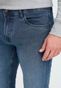 Tumedat sinised teksapüksid, slim fit lõikega, millel on ees taskud, diskreetne õmblus ja sile tekstuur. Käsi toetub kangale.