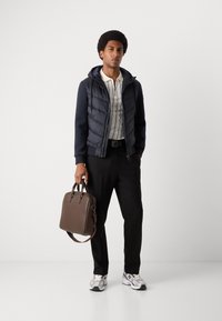 Navy puffer vest over a striped polo shirt, black pants, and white sneakers. Holding a brown leather bag with double handles.
