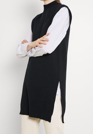 Personne portant un gilet en maille noir sans manches sur une chemise blanche à manches longues à fines rayures avec un pantalon beige, les bras croisés devant un fond uni.
