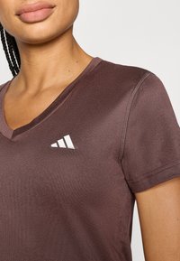 Καφέ αθλητική κοντομάνικη μπλούζα με V-λαιμόκοψη, με λευκό λογότυπο στο στήθος, κατασκευασμένη από λείο, ελαστικό ύφασμα.