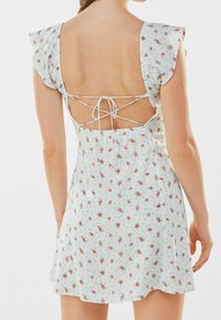 Valkoinen kukkamekko, jossa on selkäaukko, elastinen vyötärö ja lyhyet röyhelöhihat, joissa on punaisia ja vihreitä kukkakuoseja.