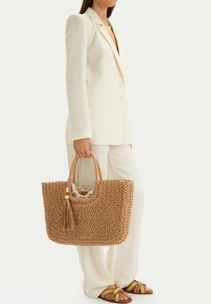 Mujer con traje crema sosteniendo un bolso grande de paja tejido con borla, usando sandalias a juego tejidas, de pie contra un fondo blanco.