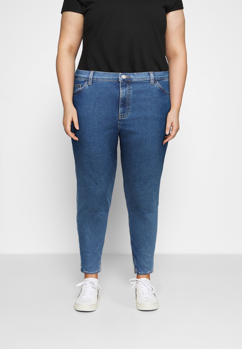 Person iført blå skinny jeans med højt talje, en sort kortærmet skjorte og hvide sneakers, stående mod en ensfarvet baggrund.