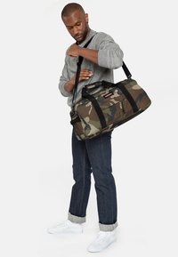 Borsa da viaggio mimetica realizzata in tessuto resistente, dotata di due manici, una tracolla e una tasca con cerniera. I colori includono tonalità di verde e marrone.