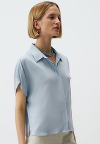 Lichtblauw shirt met korte mouwen en een kraaghalslijn, zachte textuur en zijsplitten voor meer bewegingsvrijheid. Gecombineerd met een blauwe kralenketting.