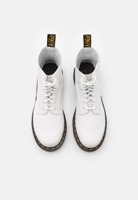 Pantofi sport înalți din piele albă cu șireturi, talpă neagră și cusături galbene. Prezintă un tab de prindere branduit în negru și galben în spate.