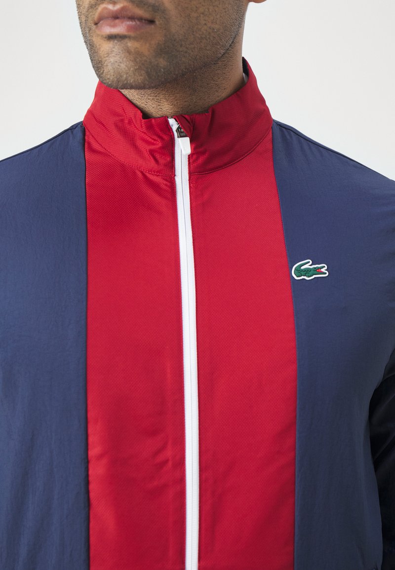 Homme portant une veste zippée bleu marine et rouge avec une fermeture éclair blanche et un logo de crocodile vert sur la poitrine, montré de la nuque jusqu'au milieu du torse.