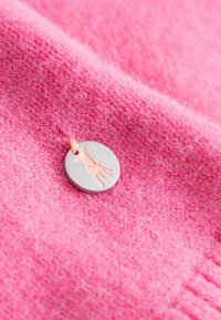 Różowy materiał polarowy o miękkiej teksturze; okrągła metalowa zawieszka z jasnoróżowym wzorem w kształcie sylwetki, przymocowana małym pomarańczowym sznurkiem.