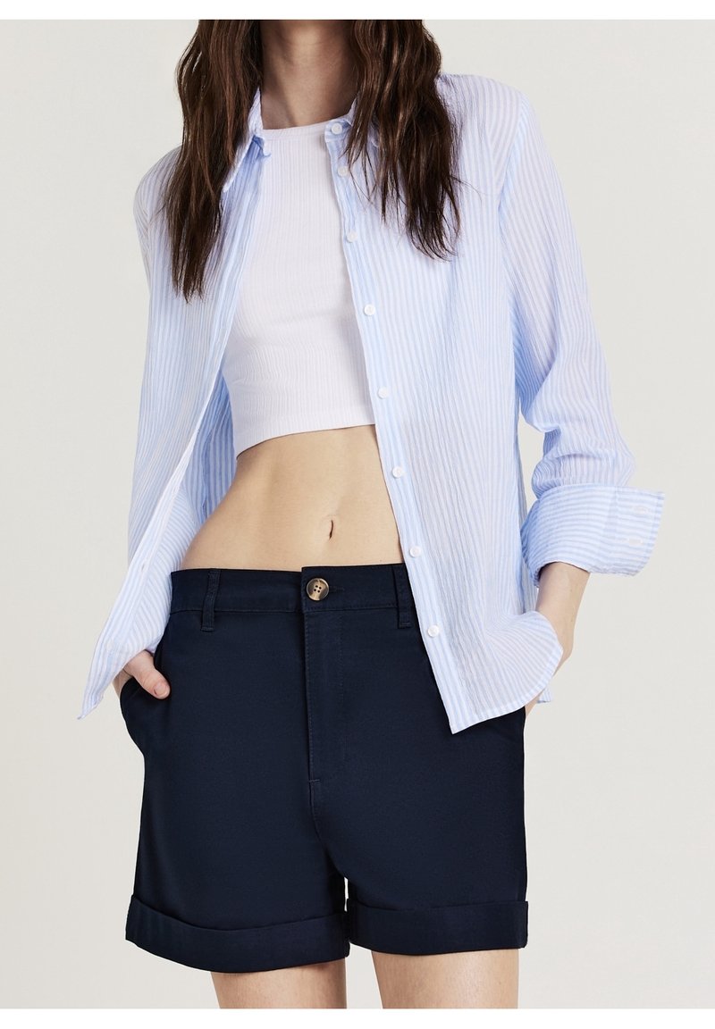 Femme portant un crop top blanc, une chemise rayée bleu clair boutonnée et un short bleu marine foncé à ourlet roulé, les mains dans les poches.