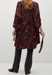 Vêtement de style kimono noir avec motif floral rouge, manches larges, noué à la taille, associé à un jean foncé et des bottines.