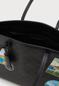 Brązowa torba na ramię z czarnymi uchwytami i wewnętrzną kieszenią na zamek, ozdobiona kolorowymi naklejkami o tematyce podróżniczej z Monako i Nowego Jorku.