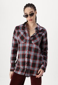 Camicia a quadri nera e grigia con accenti rossi. Maniche lunghe, chiusura con bottoni, due tasche sul petto e materiale in cotone morbido.