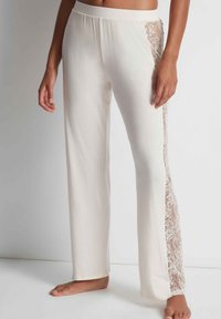 Pantalons de détente en tissu blanc cassé avec une coupe évasée, comportant des panneaux en dentelle transparente sur les côtés et une taille élastique.