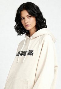 Felpa con cappuccio beige in tessuto morbido con una grande scritta nera sul davanti e coulisse. Presenta una tasca a marsupio e una vestibilità comoda.