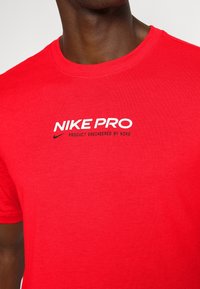 Punainen Nike Pro -t-paita, joka on valmistettu pehmeästä kankaasta. Siinä on pyöreä kaula-aukko ja musta-valkoinen painettu logo, jossa lukee "NIKE PRO" ja "PRODUCT ENGINEERED BY NIKE."