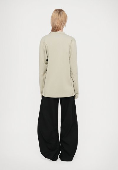 Cămașă lungă cu mânecă lungă, texturată, de culoare verde deschis, cu un croi lejer, asociată cu pantaloni negri lărgi, cu cusături vizibile și buzunare laterale. Fundal simplu.