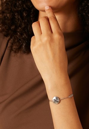 Zilveren kettingarmband met een ronde, gestructureerde bedel met ingewikkelde ontwerpen. De ketting heeft een logo-accent nabij de sluiting.