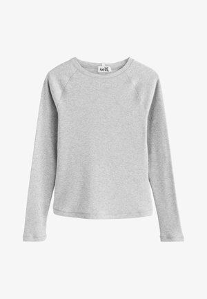 Mladá žena s kučeravými vlasmi sa usmieva, má na sebe svetlosivý dlhý rukáv a ružové nohavice, stojí vonku na slnku.