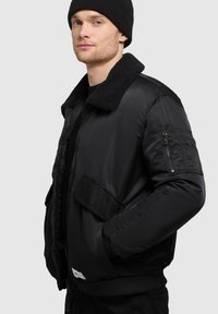 Sort bomberjakke med et shearling-krave, lynl�åslomme på ærmet og ribbede manchetter. Glat yderside med let isolering.